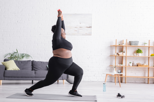 Curvy woman practicing yoga at home in black workout clothes, stretching and smiling in a well-lit room with a modern, minimalist design.